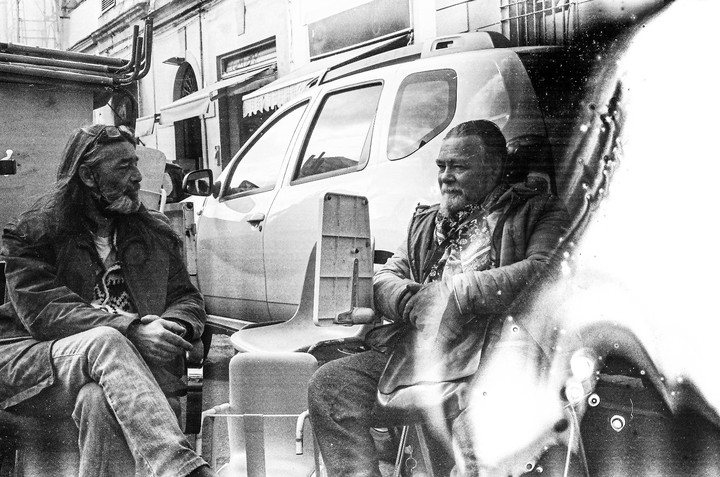 Two men sit on a street next to a car, with a large light leak on the right side of the image.