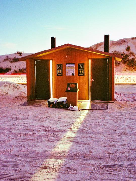 A small wooden building with two open doors and bright light inside stands on a sandy ground with tire tracks, with sand dunes in the background.