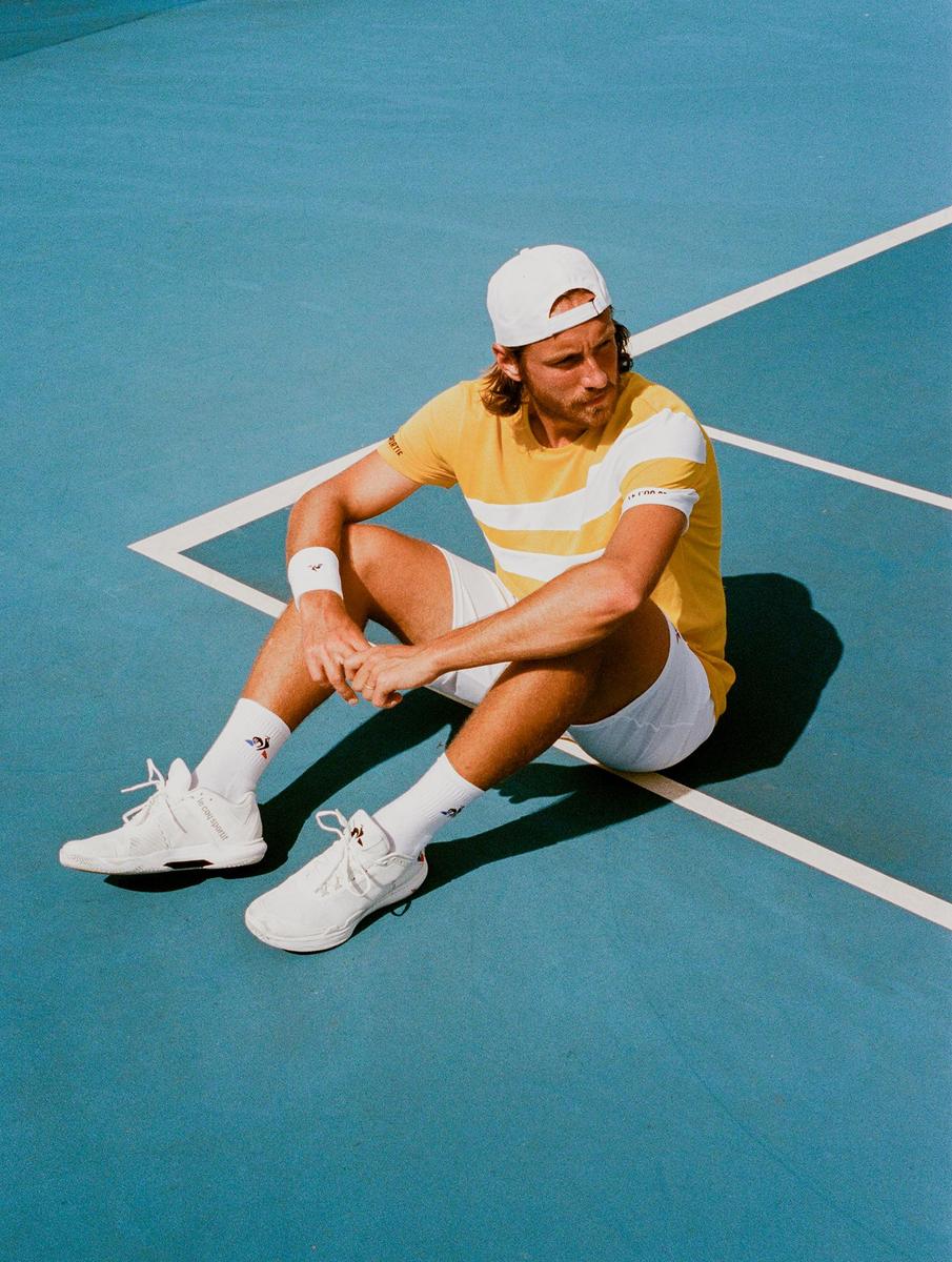 A man in a yellow shirt, white shorts, and a white cap sits on a blue tennis court.