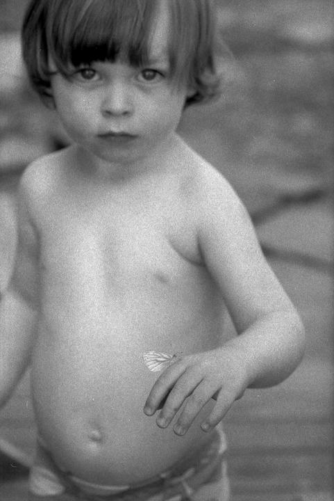 A child with short hair and bangs looks directly at the viewer, with a butterfly resting on their stomach.