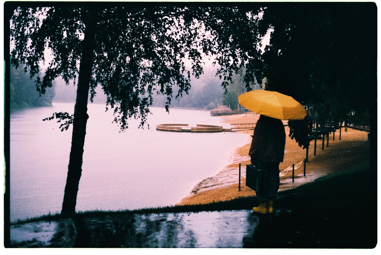 A person with a yellow umbrella stands by a lake with a circular dock on a rainy day.