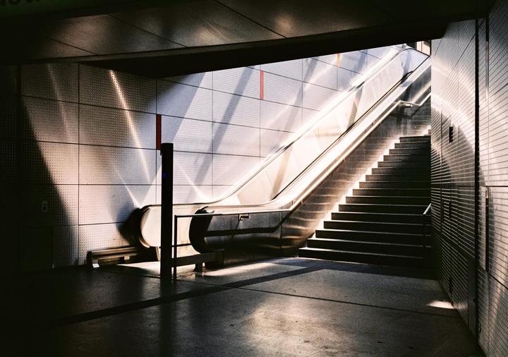 Sunlight casts strong shadows on an escalator and staircase in a subway station.