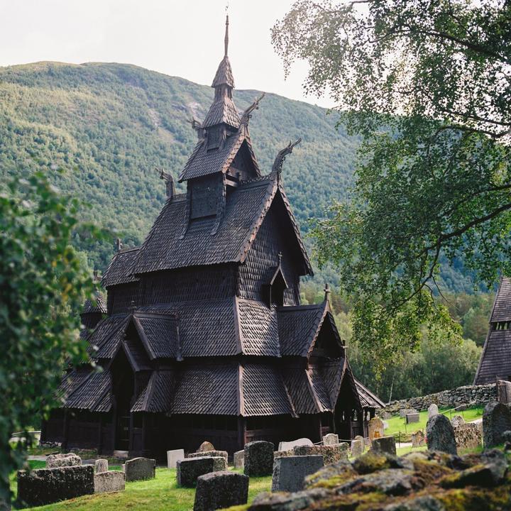 A dark wooden stave church with multiple tiered roofs stands in a graveyard with a forested mountain in the background.