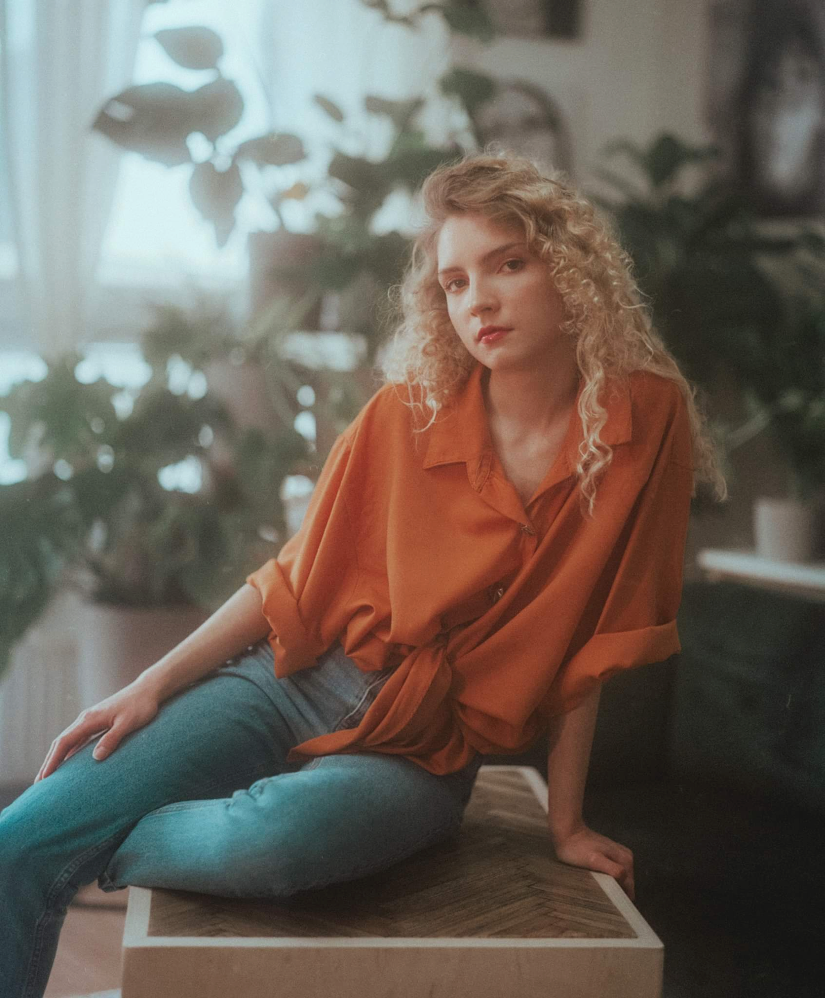 A woman with curly hair wearing an orange shirt and blue jeans sits on a wooden surface surrounded by plants.