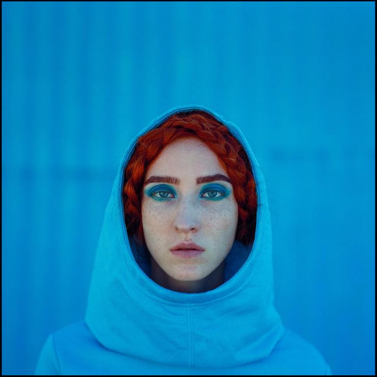 A woman with red braided hair and blue eye makeup looks directly at the viewer while wearing a blue hooded sweatshirt against a blue background.