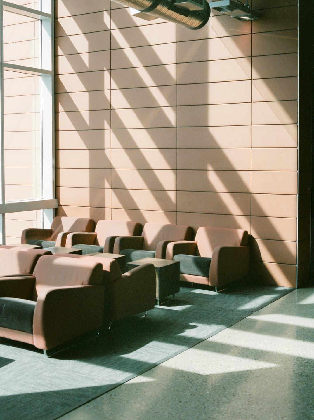 Sunlight streams through a large window, casting striped shadows across a wall and a row of armchairs in a waiting area.