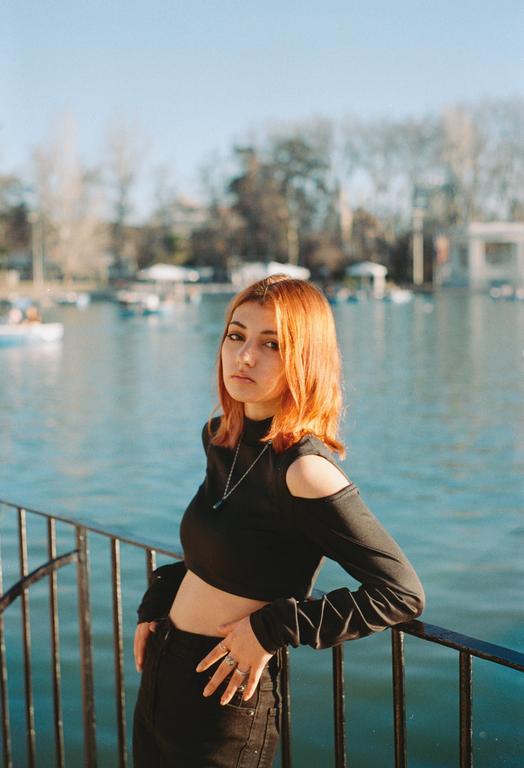 A woman with orange hair and a black top leans on a railing next to a body of water with boats.