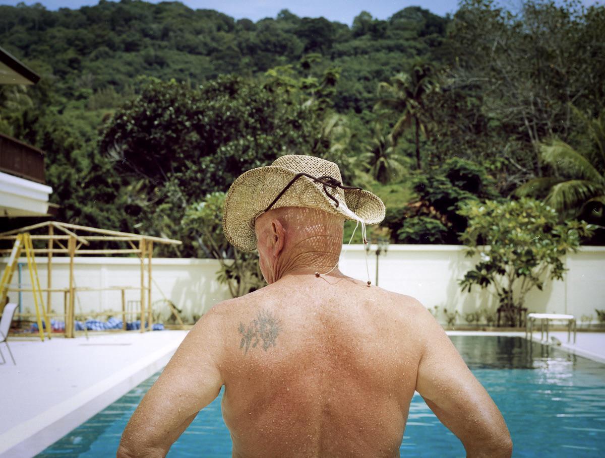 A man wearing a straw hat stands with his back to the camera, looking out over a swimming pool towards a lush green hillside.