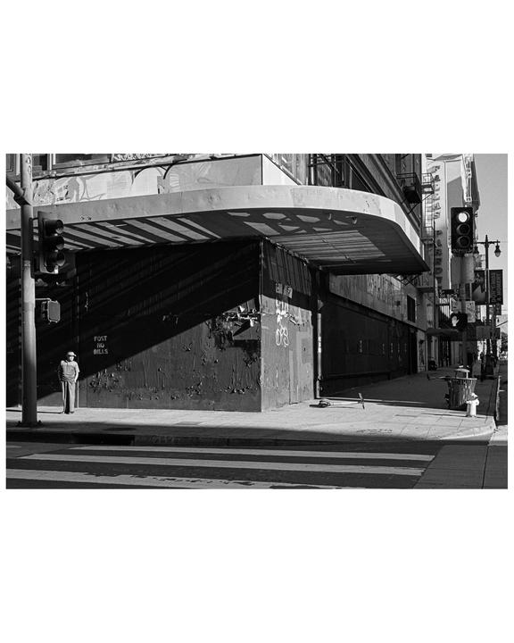 A man stands on a street corner next to a building with a curved awning and graffiti.