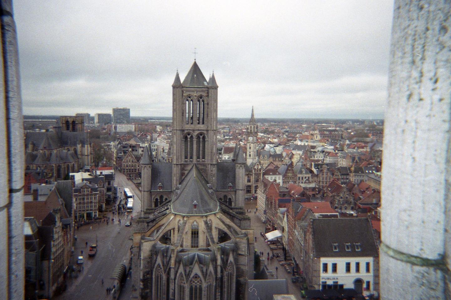 A large gothic cathedral with a tall bell tower stands in the center of a city with many buildings and a street with vehicles and pedestrians.