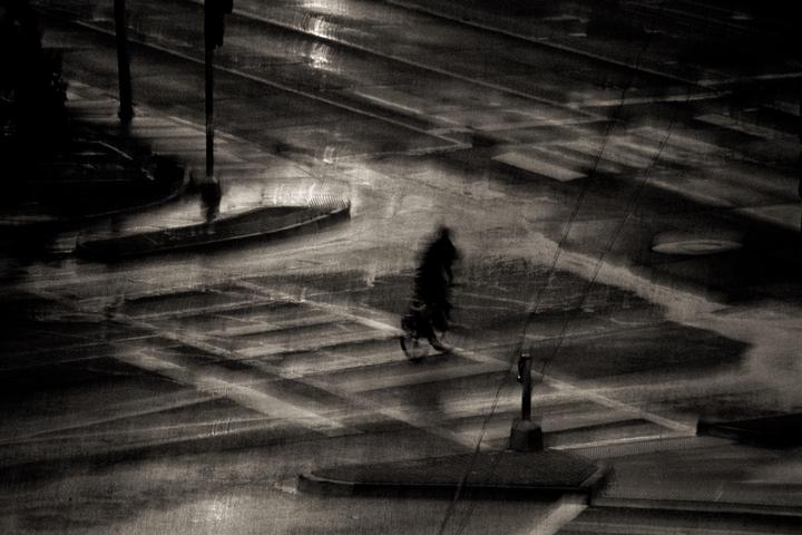 A blurry figure walks across a wet street at night, with reflections of light on the ground.