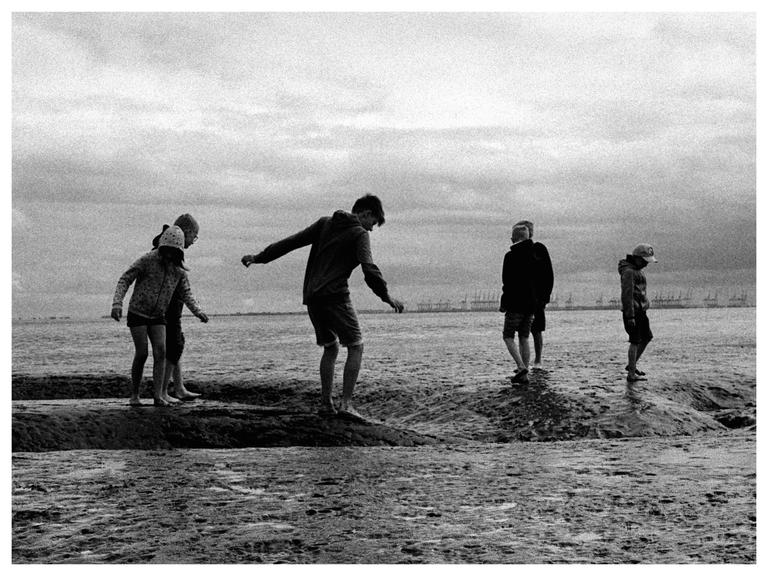 Five children walk barefoot on a rocky beach under a cloudy sky.