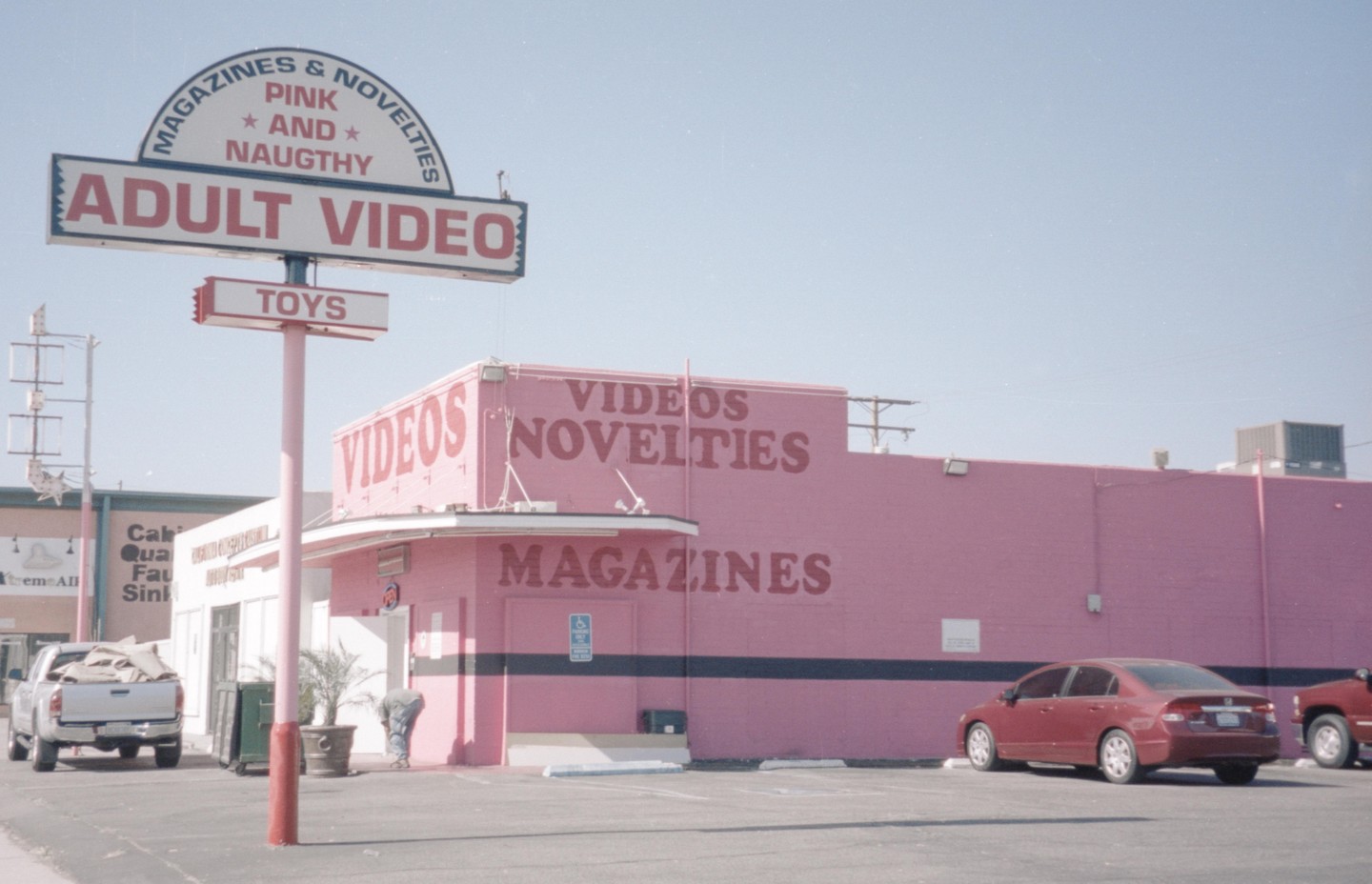 A pink building with an "Adult Video" sign stands next to a parking lot with several cars.