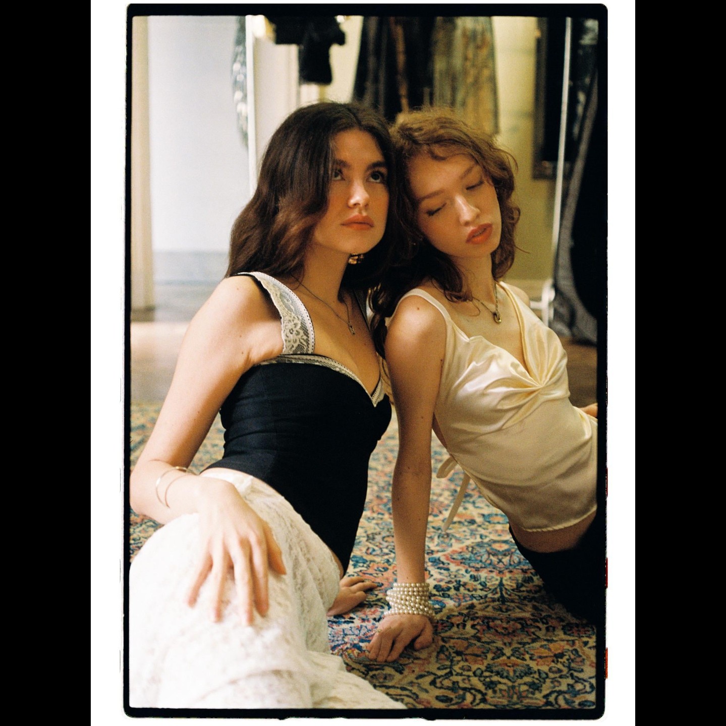 Two women are sitting on a patterned rug, one looking up and the other with her eyes closed.