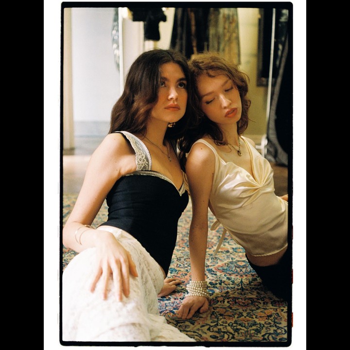 Two women are sitting on a patterned rug, one looking up and the other with her eyes closed.
