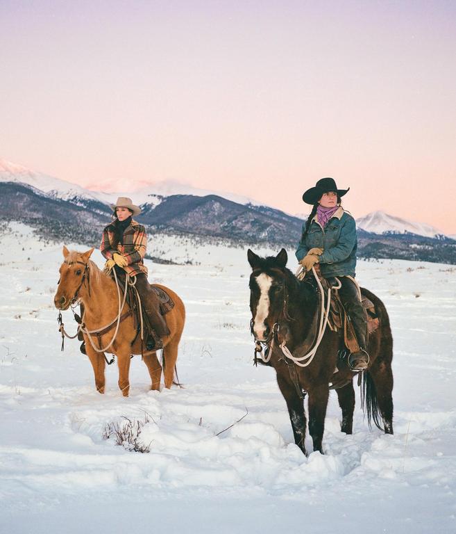 Two women on horseback stand in a snowy field with mountains in the background.