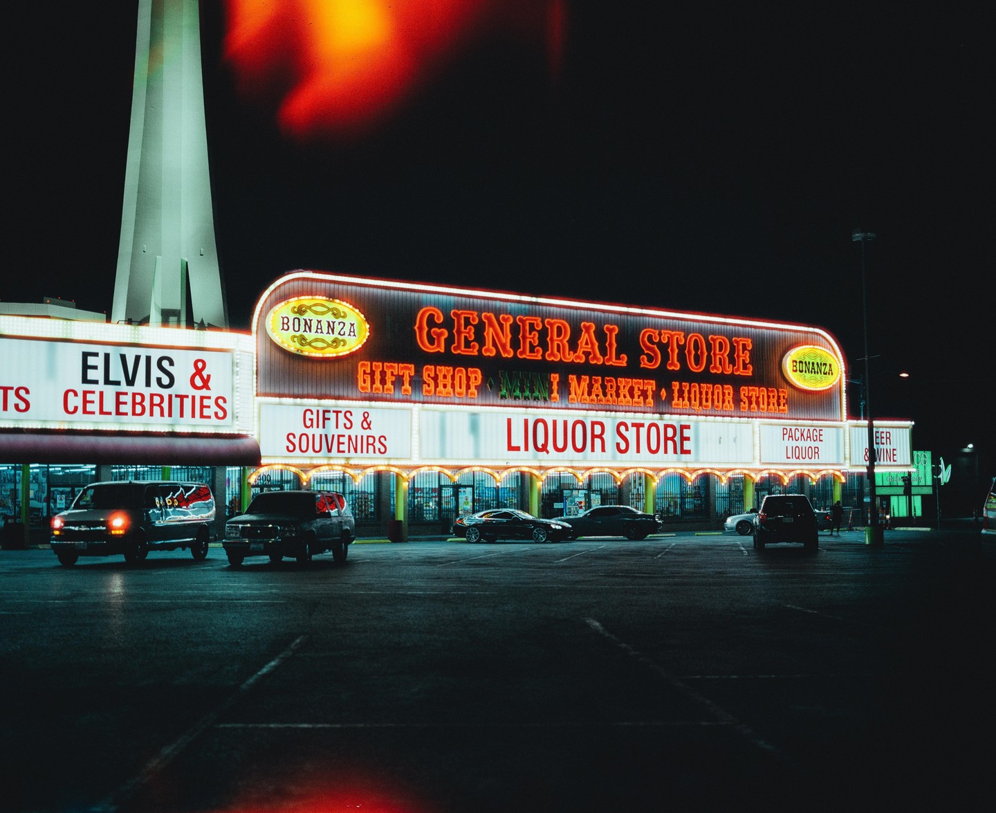 A general store with neon signs for a liquor store and gifts and souvenirs is illuminated at night with several cars parked in front.