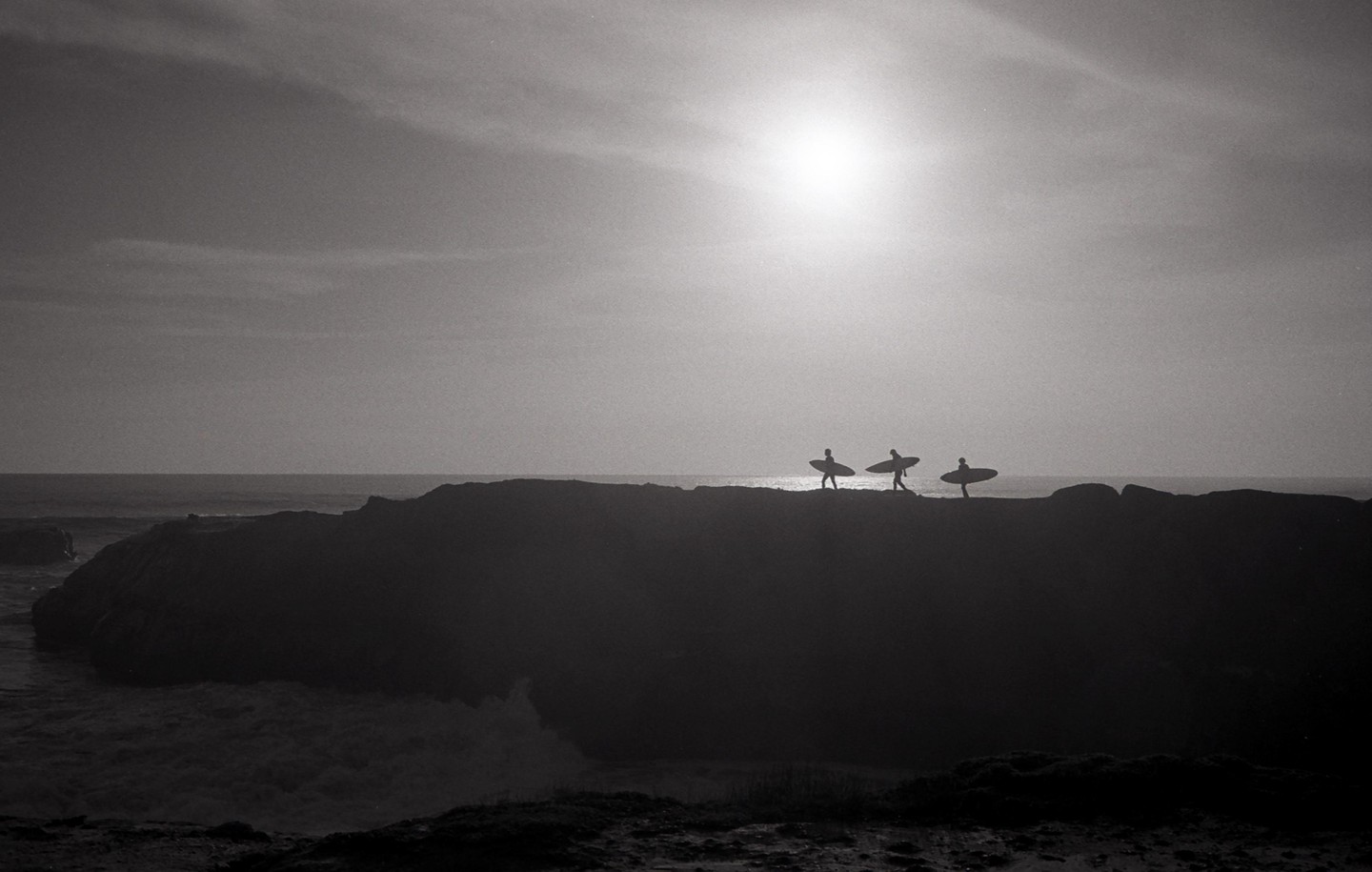 Three surfers walk along a rocky cliff edge with surfboards silhouetted against a bright sky and ocean.