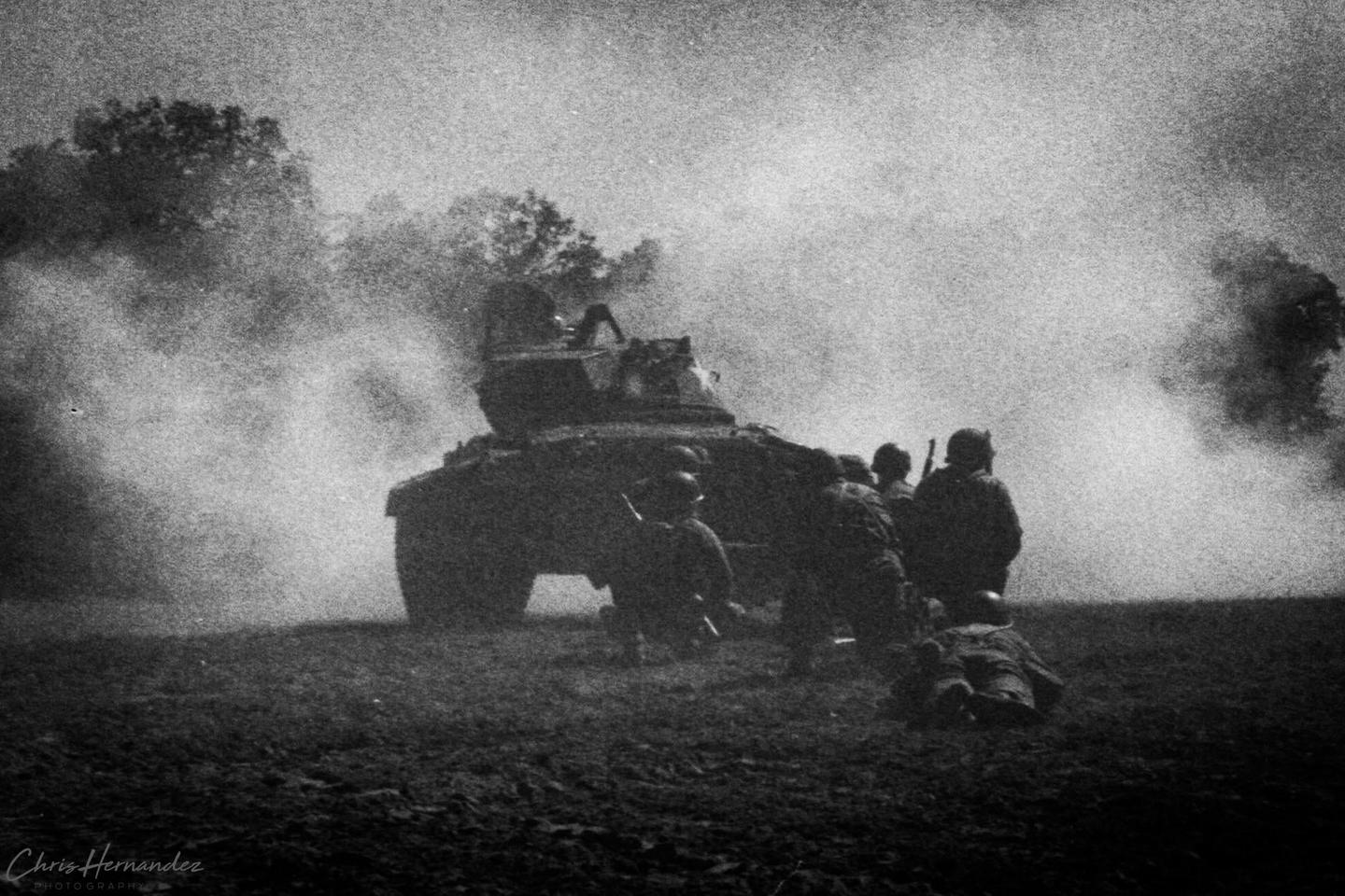 A group of soldiers are gathered around a tank on a field with smoke in the background.