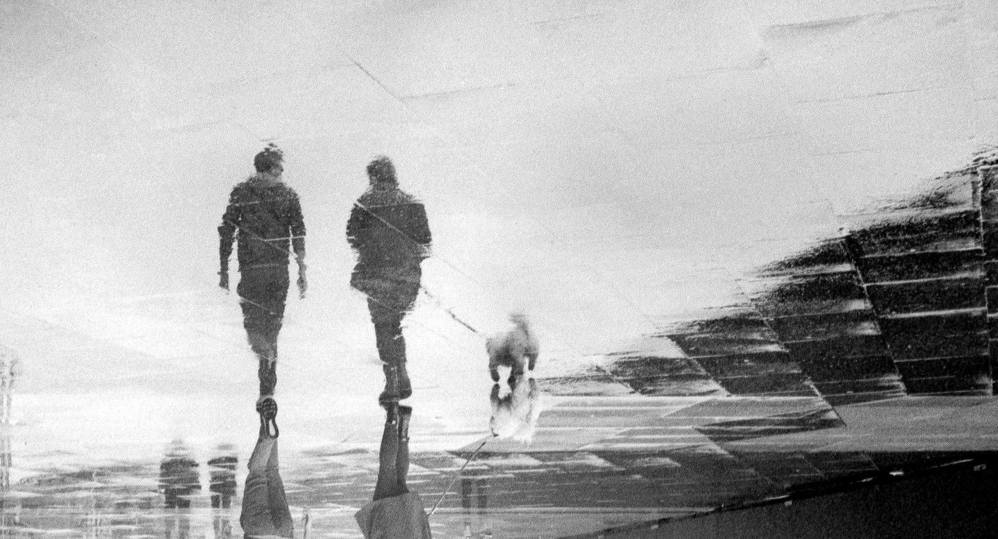Two people walk a dog on a leash, reflected in a wet surface.