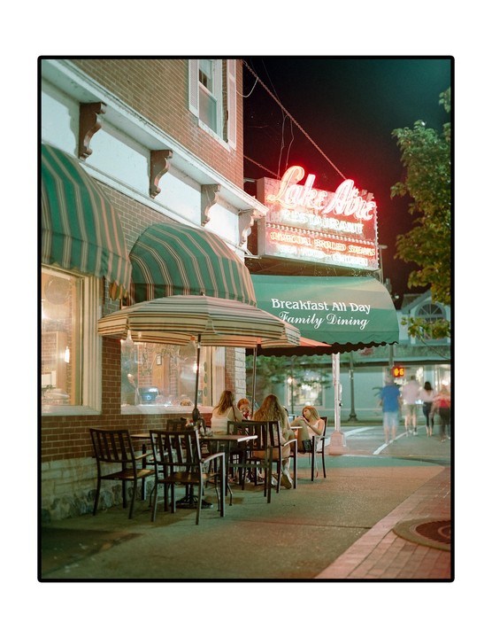 A group of people sit at outdoor tables in front of a restaurant with a neon sign at night.