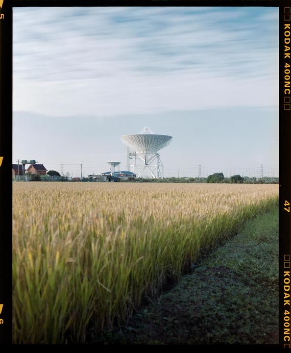 A large satellite dish stands behind a field of tall grass under a cloudy sky.