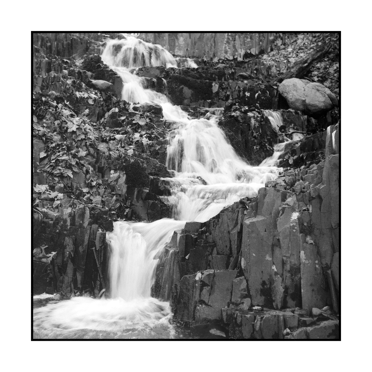 A multi-tiered waterfall cascades over dark, jagged rocks covered in foliage.