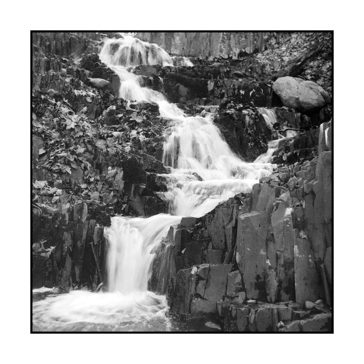 A multi-tiered waterfall cascades over dark, jagged rocks covered in foliage.