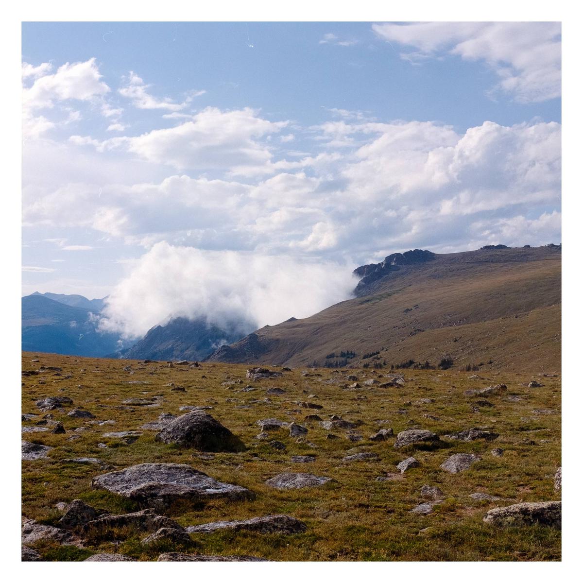Clouds roll over a mountain range with a rocky, grassy foreground.
