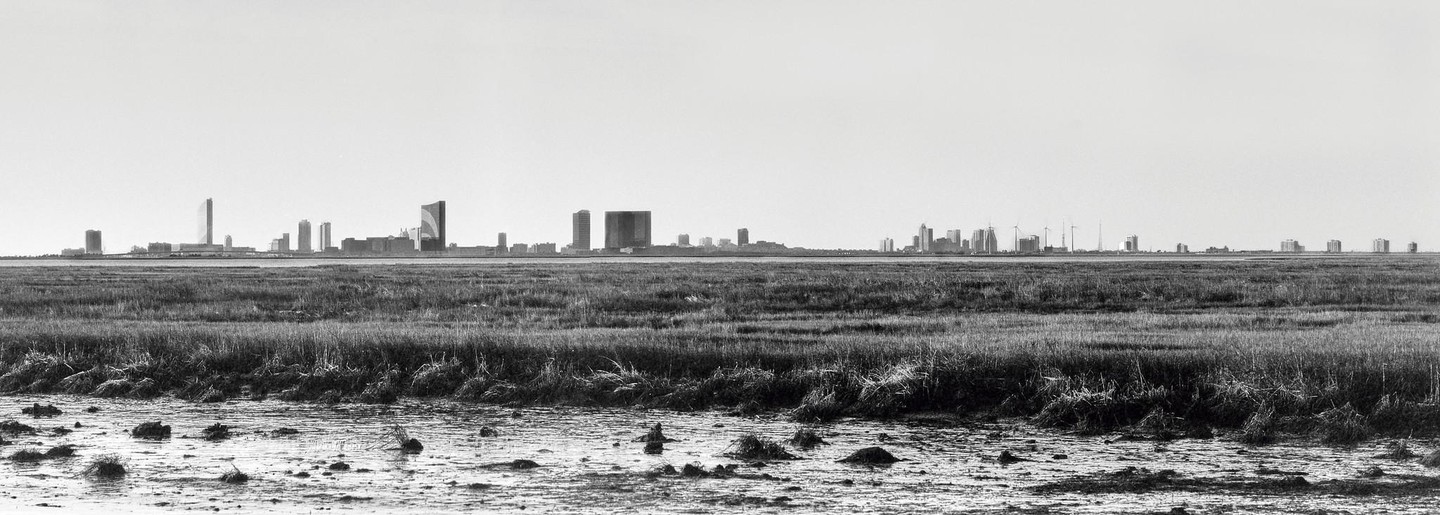 A panoramic black and white landscape shows the skyline of Atlantic City in the distance, viewed from across a vast expanse of wetlands with marsh grass and muddy water in the foreground.