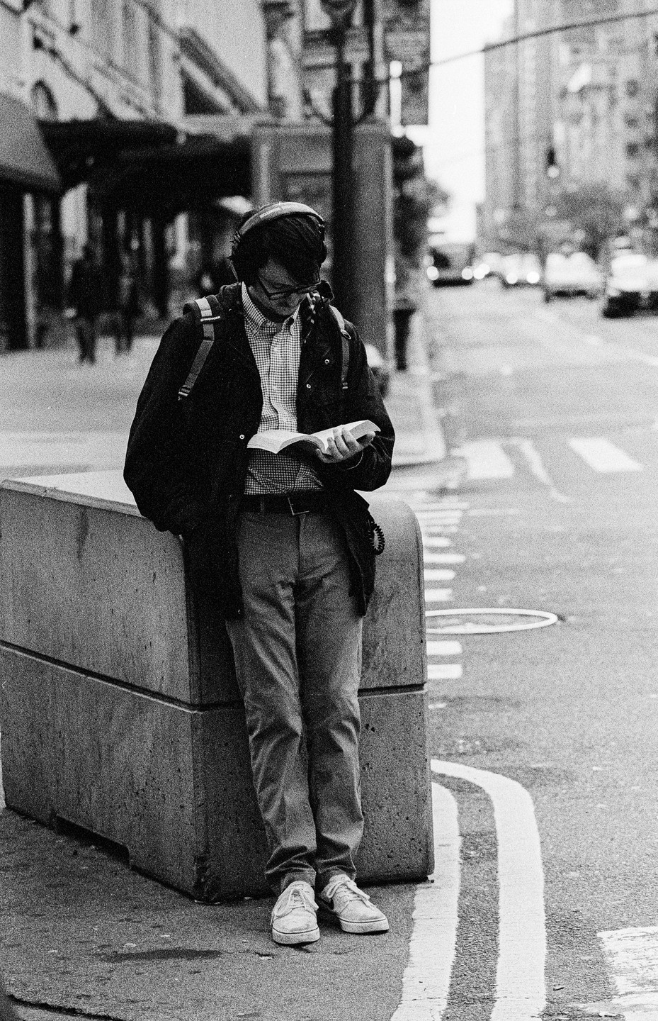 A man wearing headphones and a backpack leans against a concrete block on a city street while reading a book.