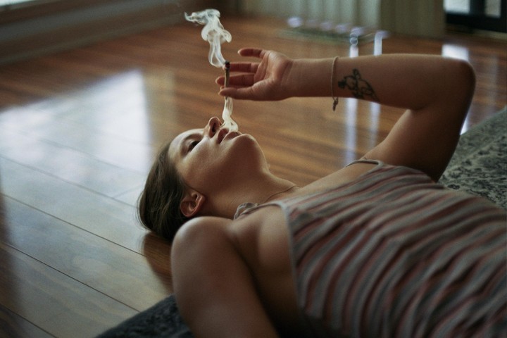 A woman lies on a wooden floor, holding a lit cigarette with smoke rising from it.