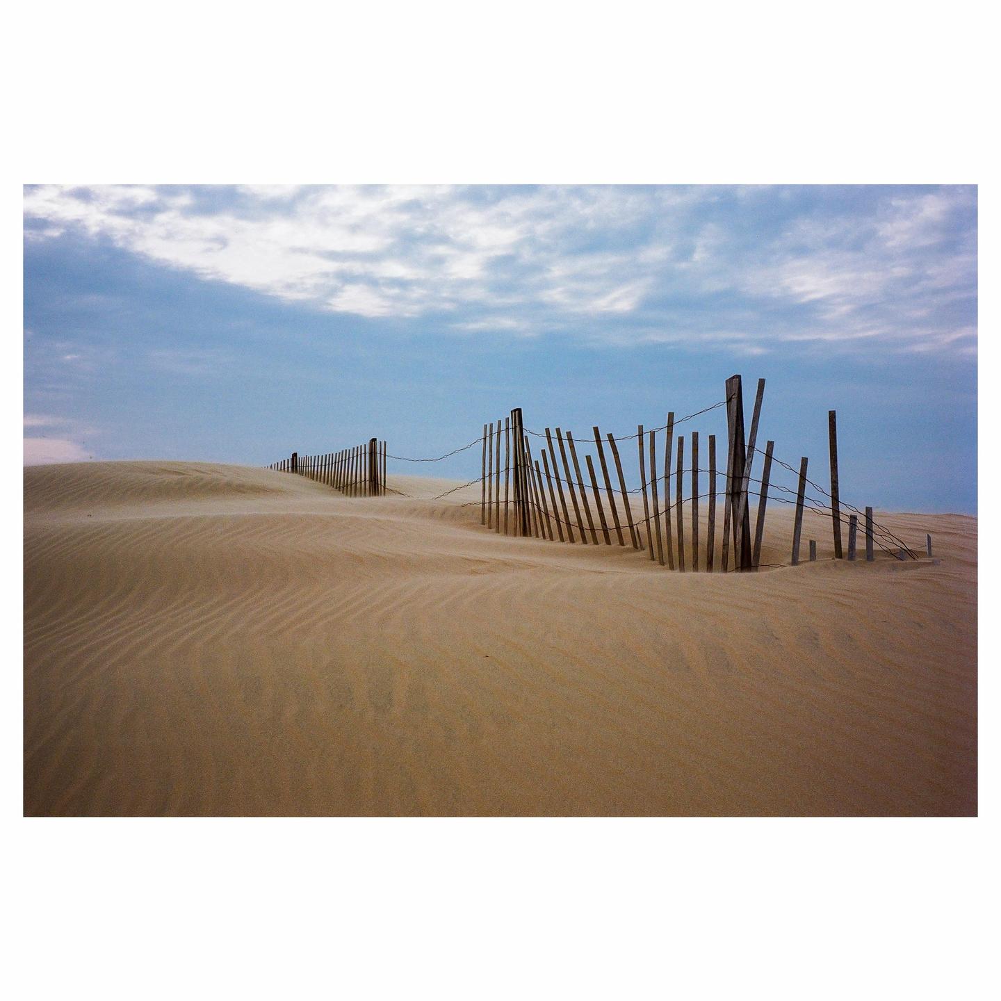 A wooden fence with wire runs across a sandy dune under a cloudy sky.