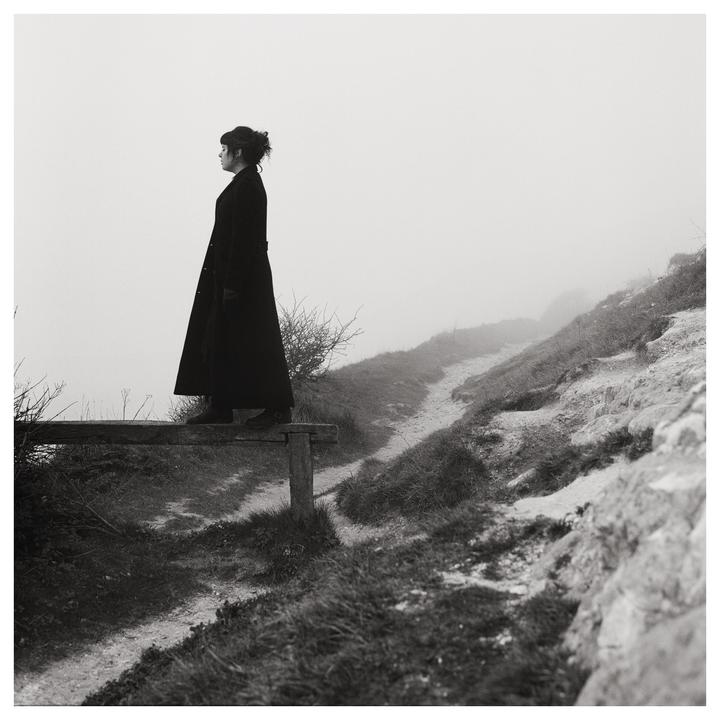 A woman in a long coat stands on a wooden barrier looking out into a foggy landscape with a path winding up a hill.