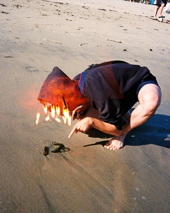 A person with a flaming head crouches on a sandy beach, pointing at something on the ground.