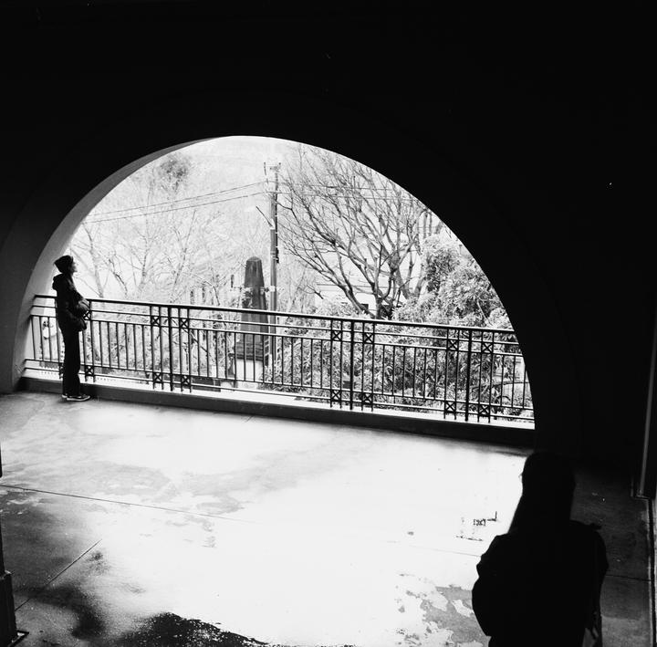 A person stands on a balcony looking out through an archway at a winter landscape while another person stands in the foreground with their back to the viewer.