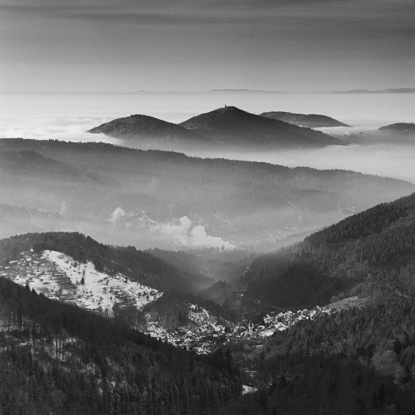 A village nestled in a valley is surrounded by forested mountains, with a sea of fog in the distance.