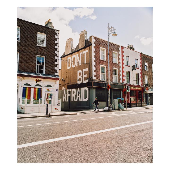 A person walks on a sidewalk next to a building with "DON'T BE AFRAID" painted on its side.