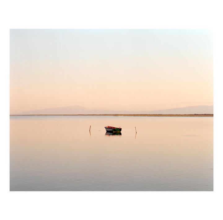 A small boat floats on calm water with mountains in the distance under a pale sky.