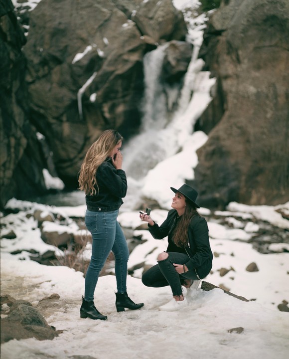 A woman kneels in the snow to propose to another woman in front of a waterfall.
