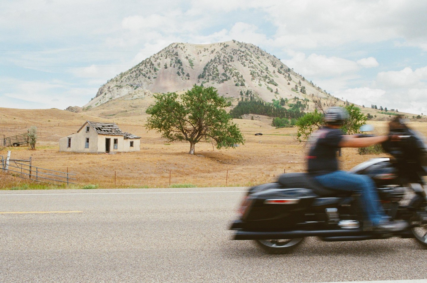 A motorcycle speeds past an abandoned house and a mountain in a dry, grassy landscape under a cloudy sky.