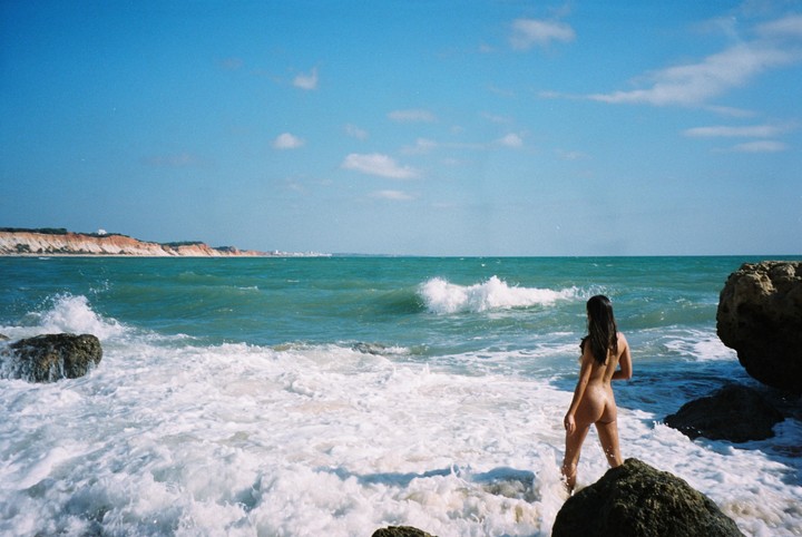 A woman stands in the foamy ocean water with cliffs in the distance under a blue sky.