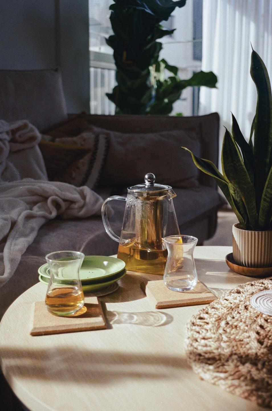 A glass teapot and two glasses of tea sit on a round wooden table in a sunlit room.