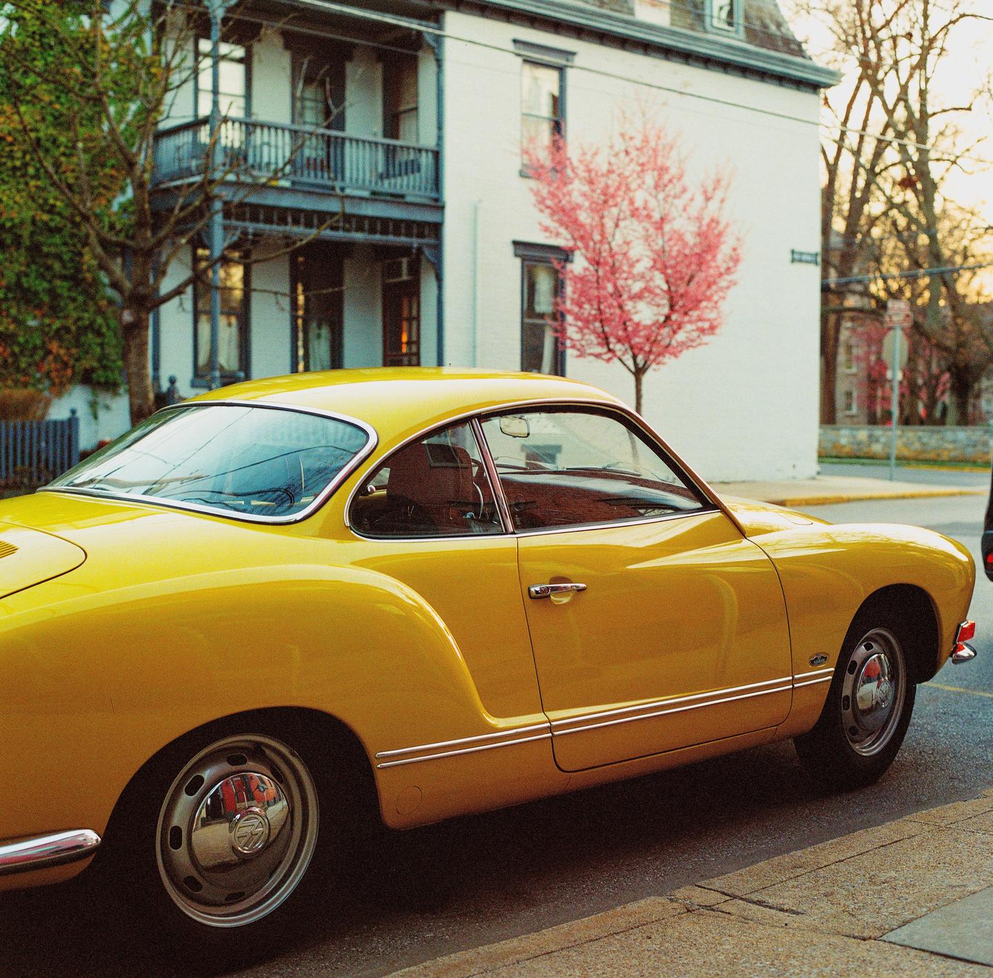 A yellow vintage car is parked on a street next to a white building with a pink tree in the background.
