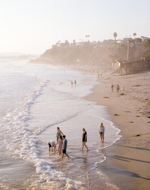 A group of people wade in the ocean at a hazy beach with a town on a hill in the background.