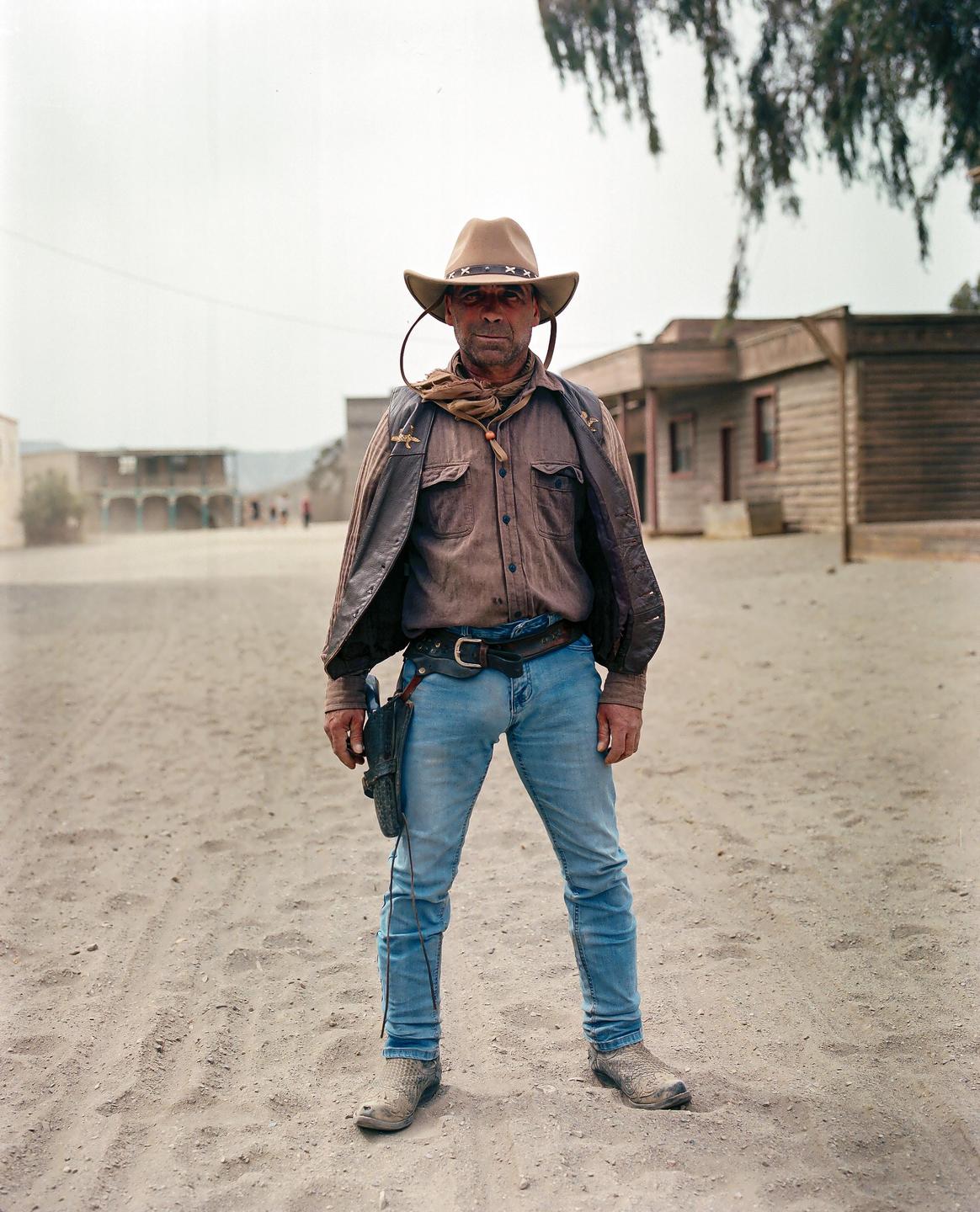 A man dressed as a cowboy stands in the middle of a dusty street in front of wooden buildings.
