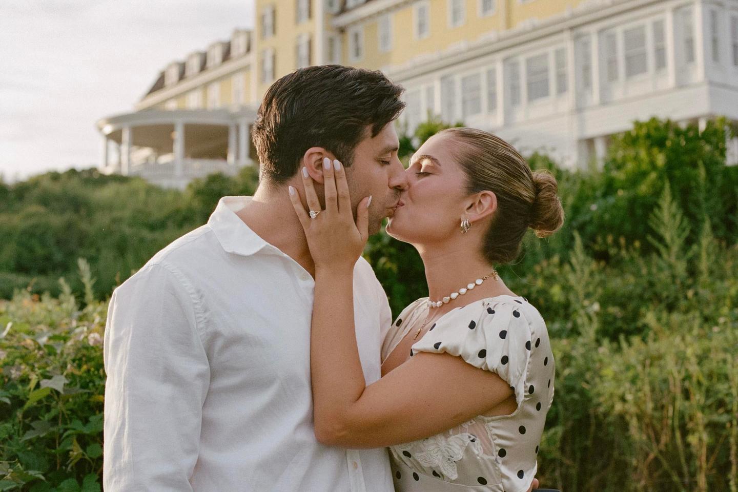 A couple kisses outdoors with a large yellow building in the background.