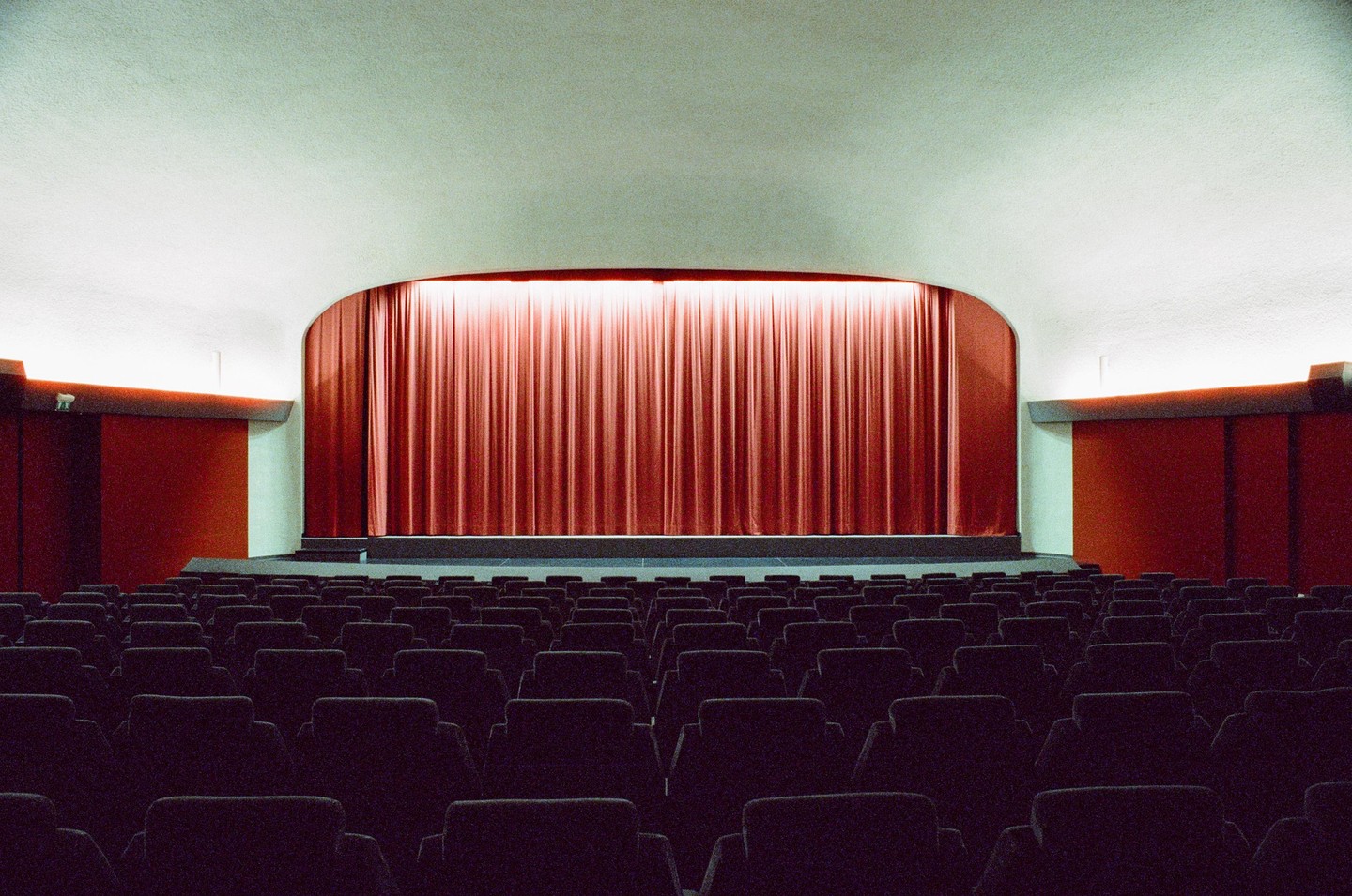 An empty theater with rows of seats faces a stage with a red curtain.