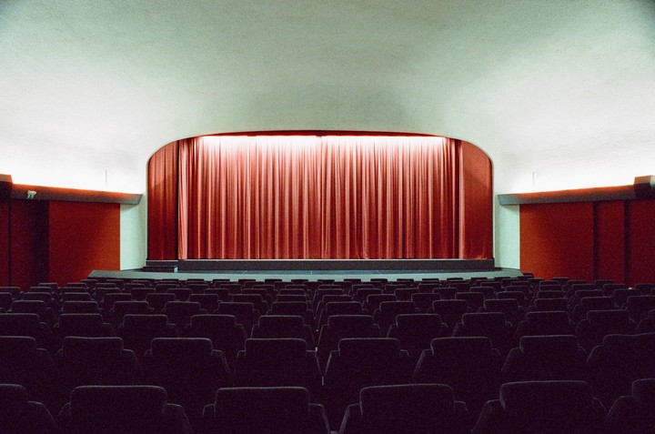 An empty theater with rows of seats faces a stage with a red curtain.