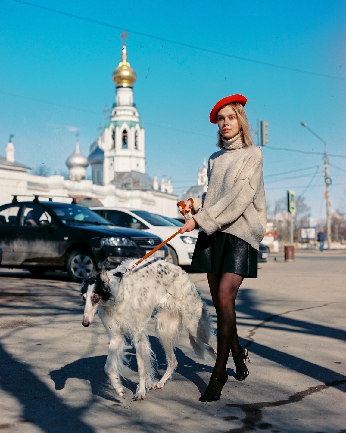 A woman in a red beret walks a borzoi dog on a leash down a street with a church in the background.
